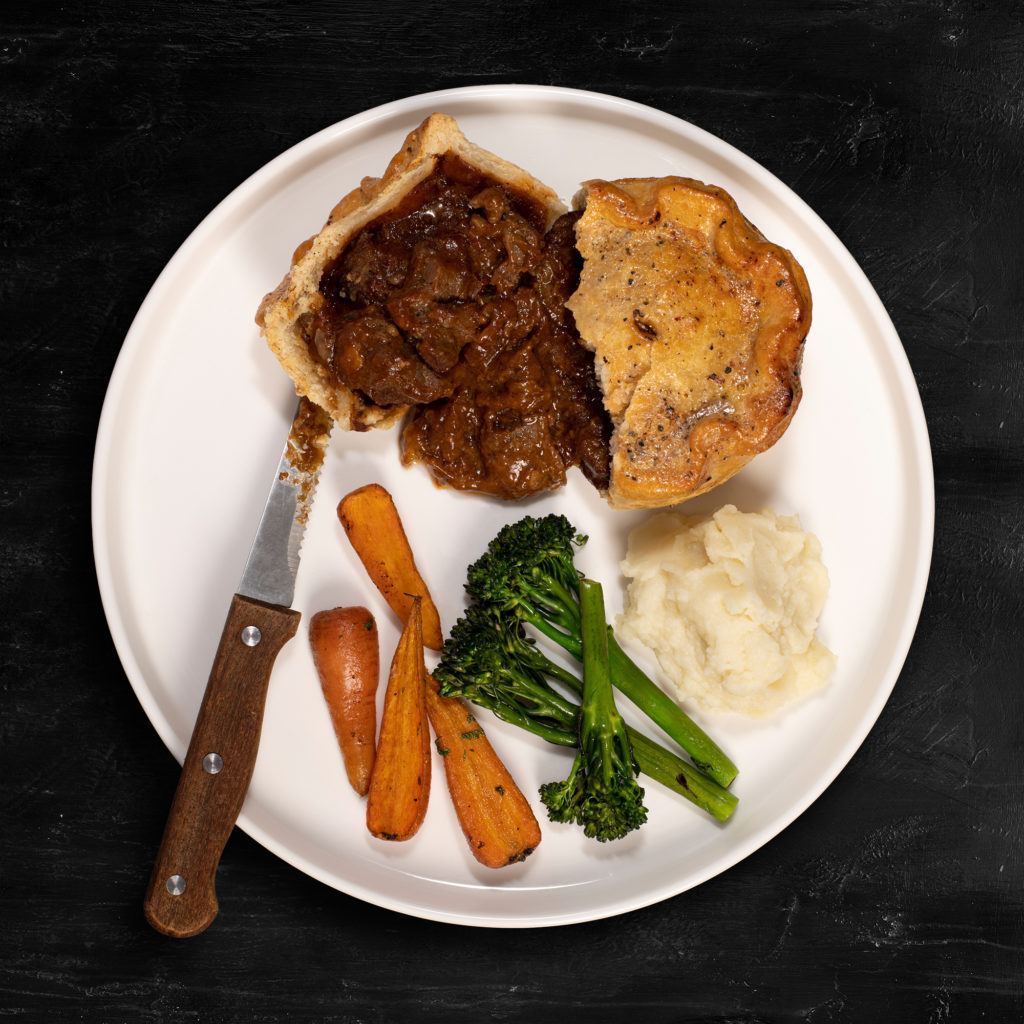 A Steak and Ale flavour pie with carrots, broccoli and mash on a plate sat on a slate background