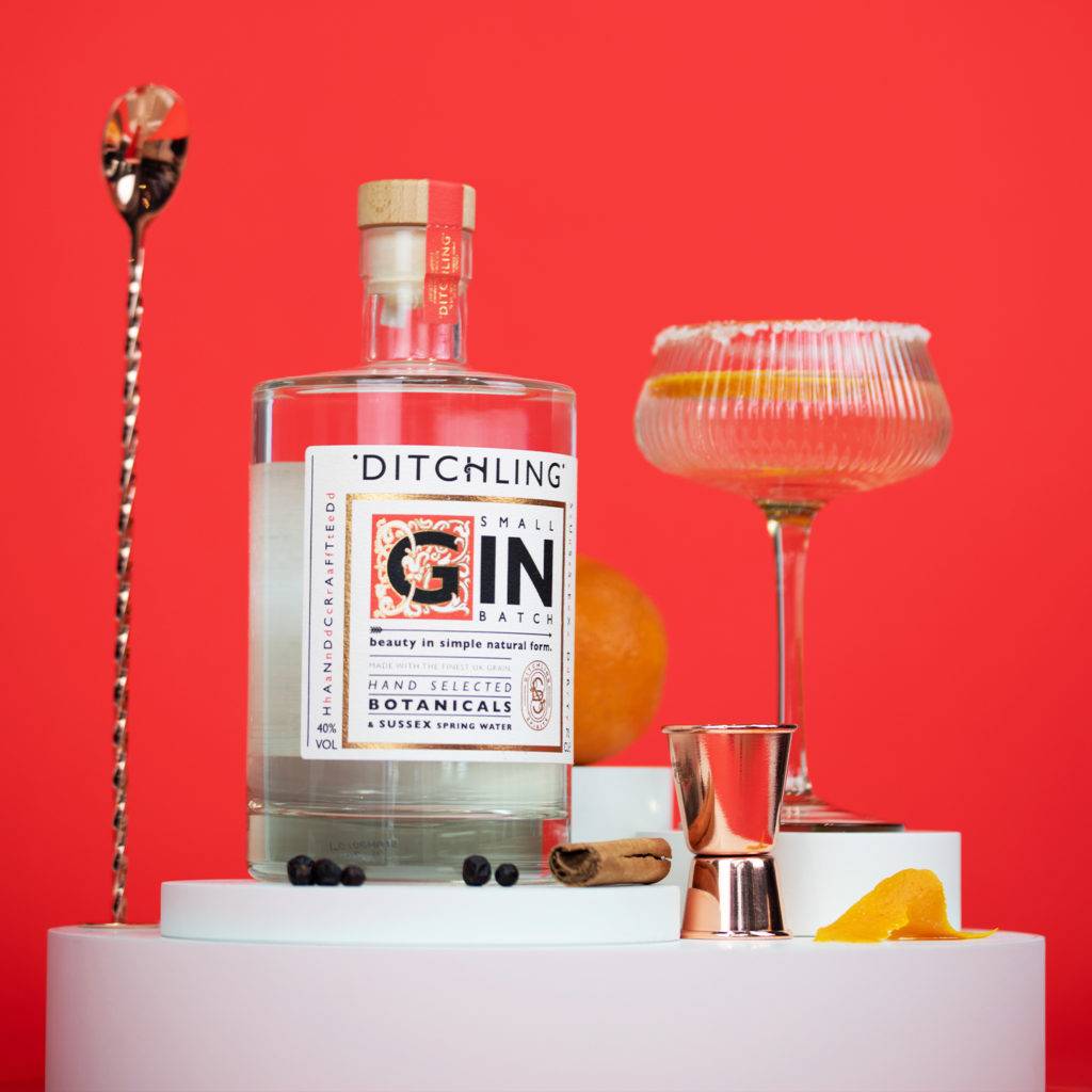 A bottle of Ditchling Gin on a bright red background, with oranges, lemon, a glass of gin with sugar around the rim and a gold stirrer