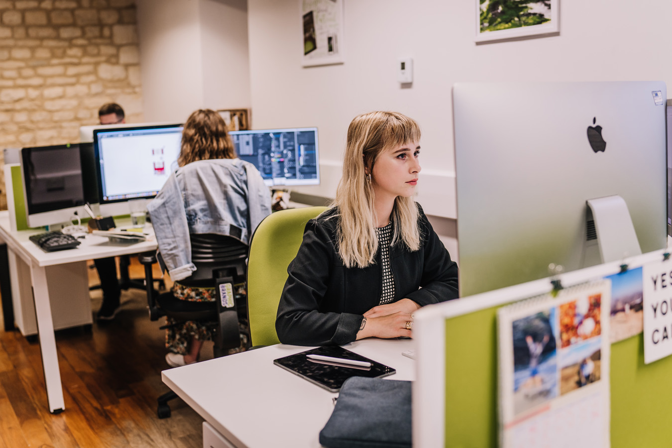 A female is sat at her desk working on an Apple PC, another female is sat hewing her working from two computer screens