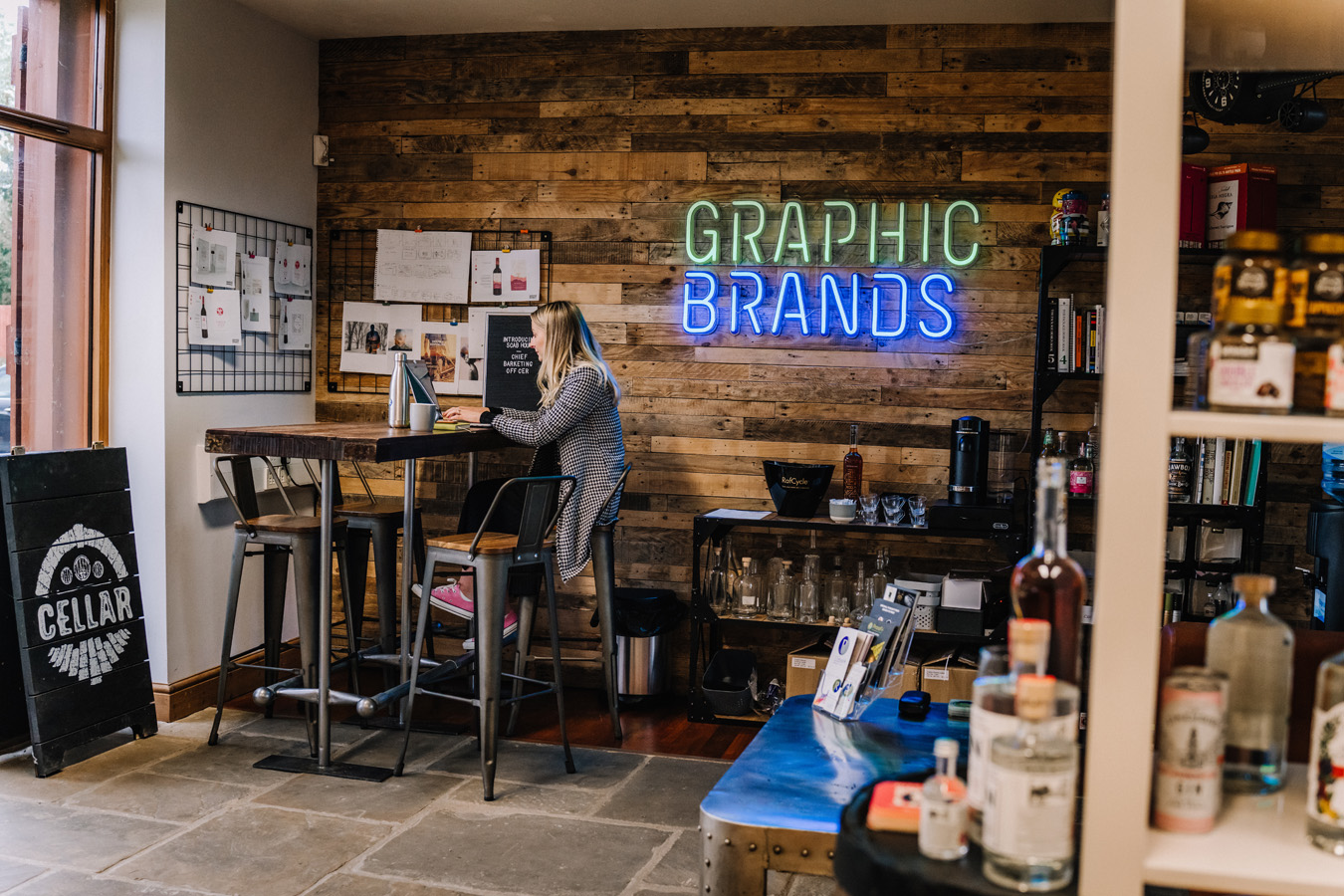 A green and blue neon sign saying Graphic Brands is hanging on a wood panelled wall in an office. A female is sitting at a table working on her laptop