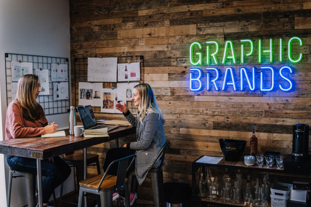 Two females sat at a desk with a blue and green sign saying Graphic Brands on the wall next to them