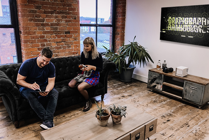 Two people sat on a black velvet Chesterfield sofa in a large spacious room with exposed brick walls and wooden floors