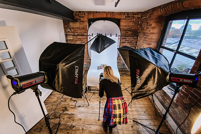 Female wearing a colourful checked skirt taking photos of a box sat on a white background. There is flash equipment around her