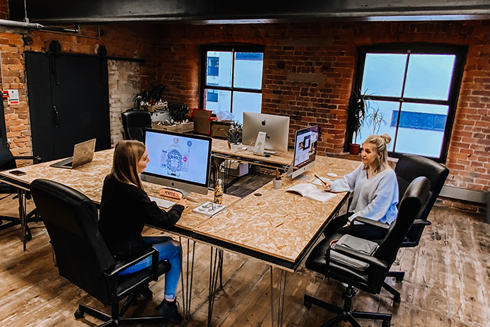 Two females sat opposite each other on a square bank of chip board desks with apple macs in front of them