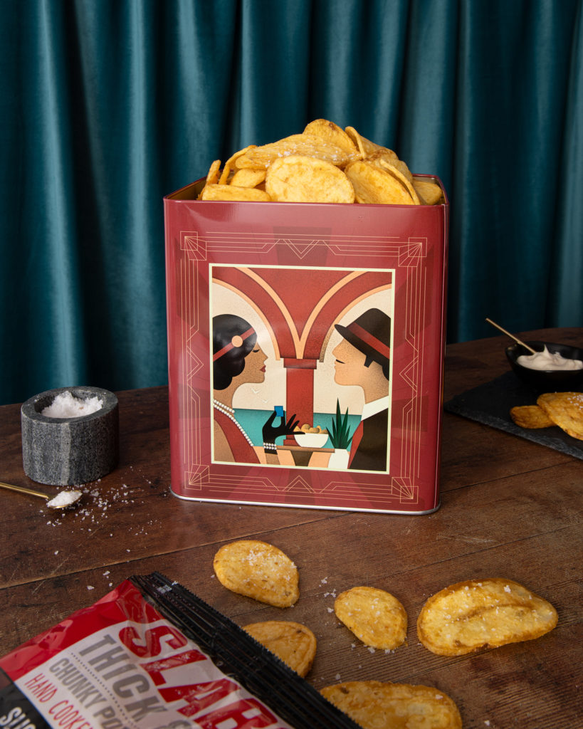 A retro looking tin filled with SLABS crisps, with an art deco inspired design with a packet of crisps and scattered crisps on the table in the foreground