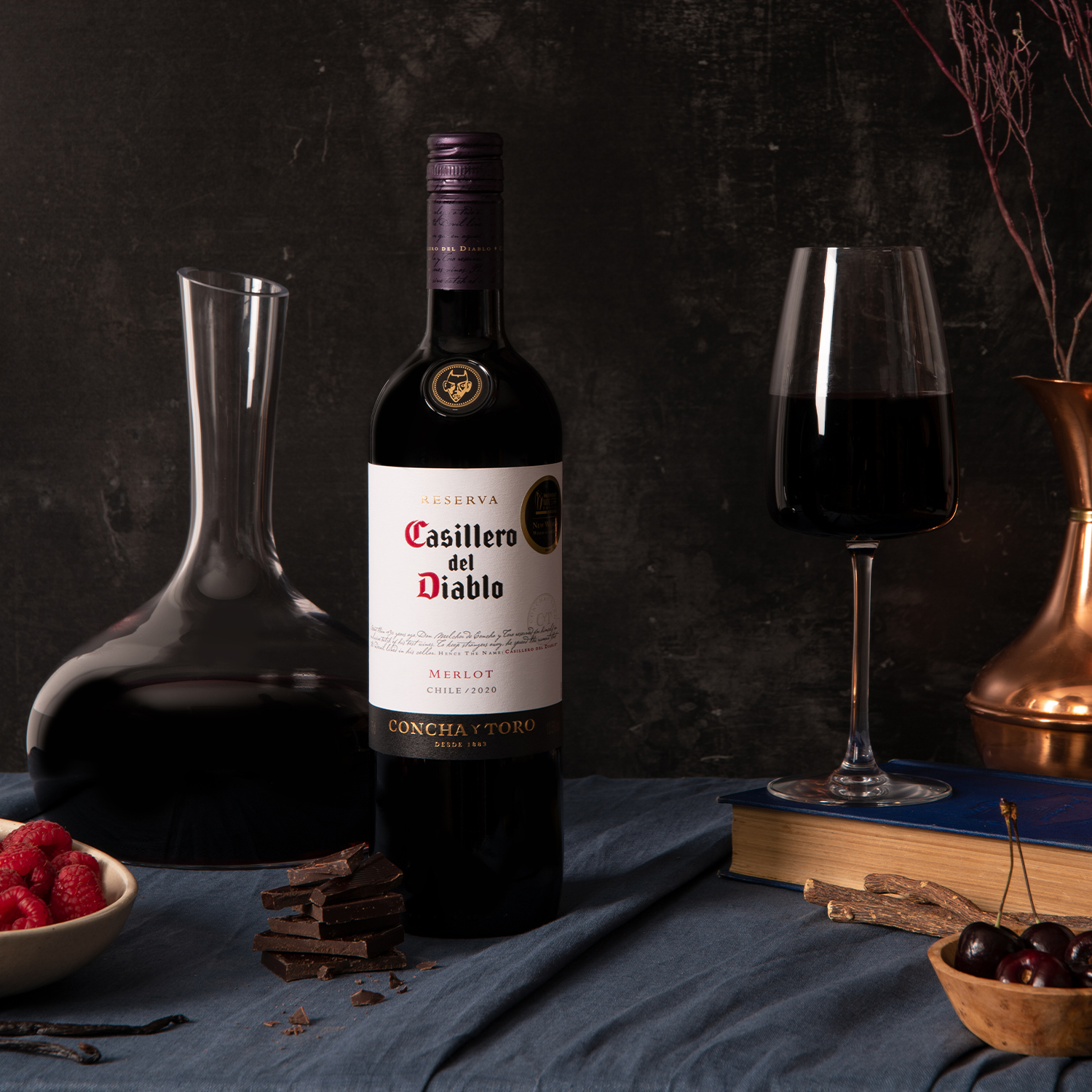 A bottle of Casillero del Diablo Merlot, styled on a dark textured background with a navy blue cloth draped on the table along side raspberries, cherries and chocolate