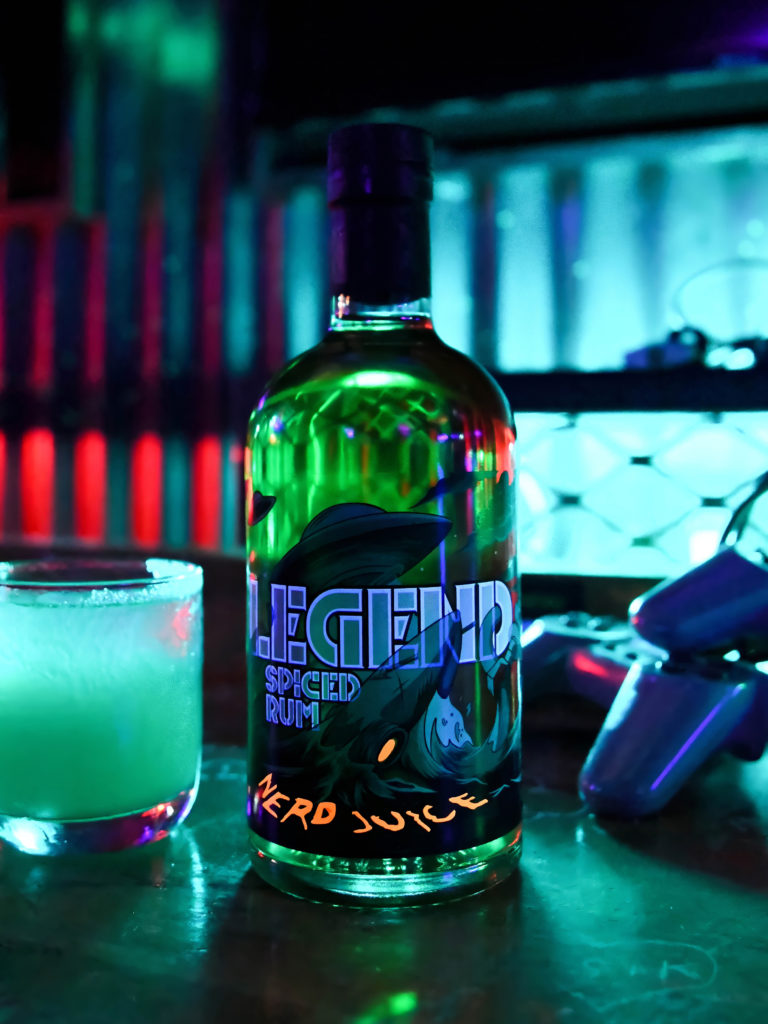 A bottle of Legend, featuring a label desng inspired by aliens and outer space, alongside a green cocktail and a pair of PlayStation controls