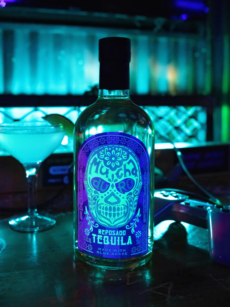 A bottle of Mucha Duro, featuring a Lucha Libre inspired label design, alongside a blue cocktail and a PlayStation control