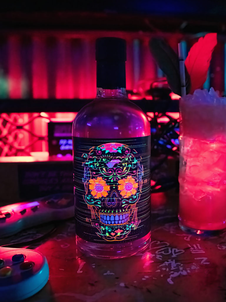 A bottle featuring our hero label design, a sugar skull inspired by Day of the Dead, made up of five UV foils, alongside a pink cocktail and a gaming control