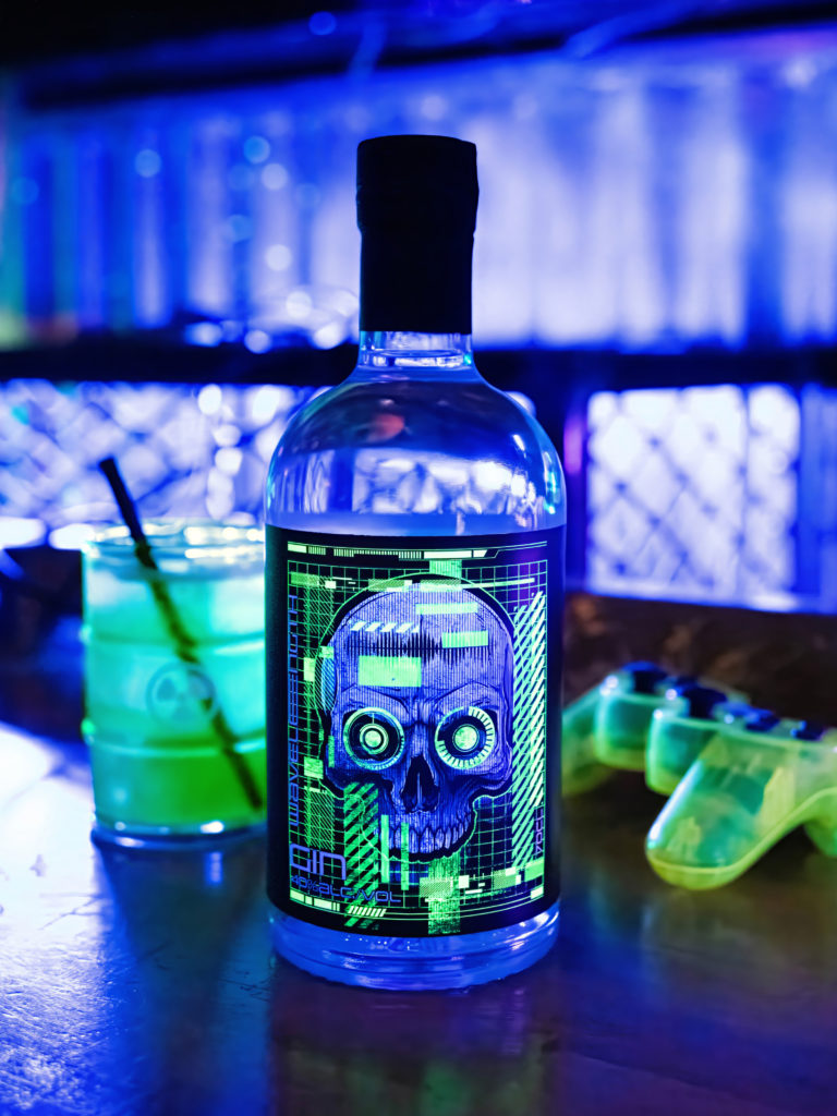 A bottle of Wavelength Gin, featuring a label design inspired by future technology and AI, alongside a bright green cocktail and neon glowing gaming control