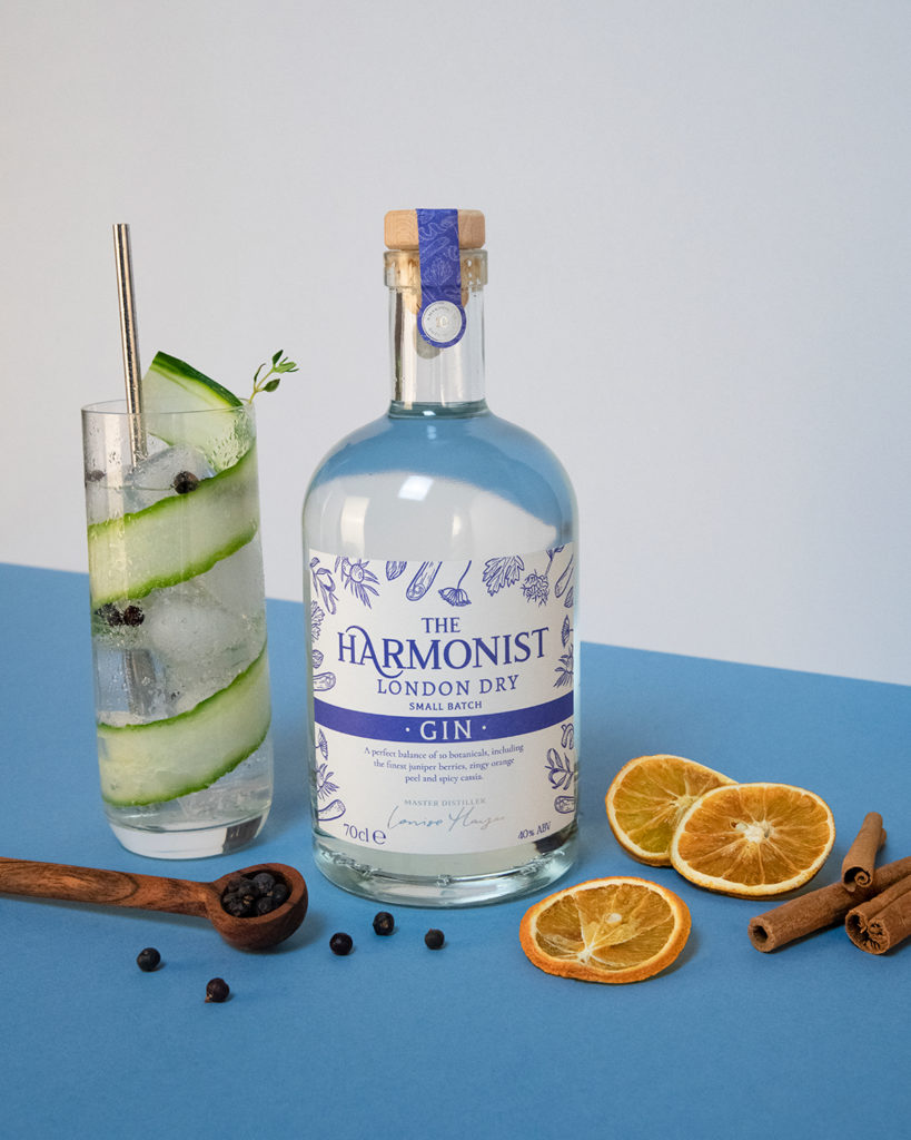 A bottle of The Harmonist London Dry Gin with a glass of gin filled with slice of cucumber, a wooden spoon, dried orange peel and sticks of cinnamon.